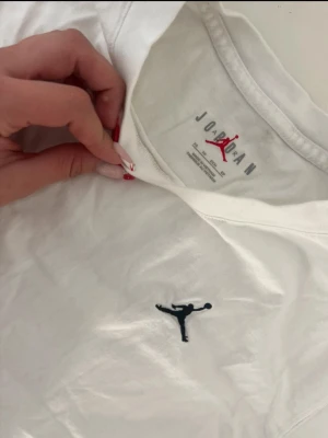 Vit Jordan t-shirt med logga - Vit t-shirt från Jordan med klassisk svart Jumpman-logga broderad på bröstet. T-shirten har rund halsringning och är tillverkad i mjuk bomull för en bekväm känsla. Perfekt för en sportig och avslappnad stil.