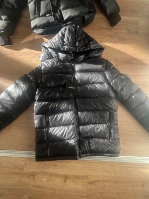 Svart dunjacka från Moncler med huva - Snygg svart dunjacka från Moncler med glansig finish och huva. Jackan har dragkedja framtill, två sidofickor med dragkedja och Moncler-logga på ärmen. Perfekt för kalla dagar och ger en cool streetstyle-look.