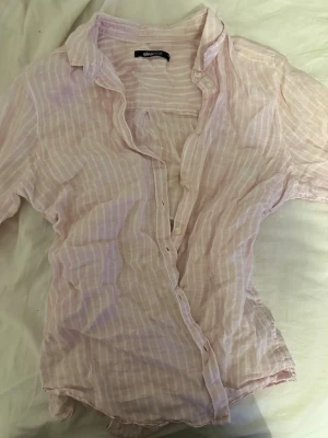 Ljusrosa randig skjorta från Gina Tricot - Luftig och tunn skjorta i ljusrosa med vita ränder från Gina Tricot. Skjortan har klassisk krage, knappar framtill och långa ärmar. Perfekt för varma dagar och enkel att styla till olika outfits.