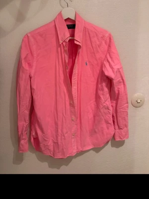 Rosa skjorta från Polo Ralph Lauren - Klassisk rosa skjorta från Polo Ralph Lauren med button-down krage och blå broderad logga på bröstet. Skjortan har långa ärmar och knäppning framtill. Perfekt för dig som vill ha en färgstark och stilren look.