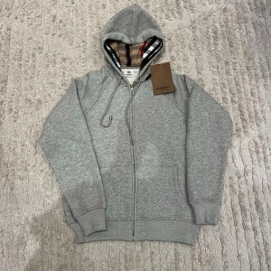 Grå zip-hoodie från Burberry - Snygg grå hoodie med dragkedja från Burberry. Klassisk huva med det ikoniska rutmönstret på insidan, ribbade muddar och fickor framtill. Perfekt för en avslappnad och stilren look.