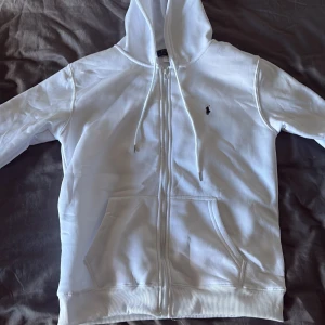 Vit hoodie från Polo Ralph Lauren L - Vit hoodie från Polo Ralph Lauren med svart broderad logga på bröstet. Tröjan har dragkedja, huva med snörning och två stora fickor framtill. Tillverkad i mjuk bomullsmix, perfekt för chill dagar. Klassisk och stilren design. Passar L med 