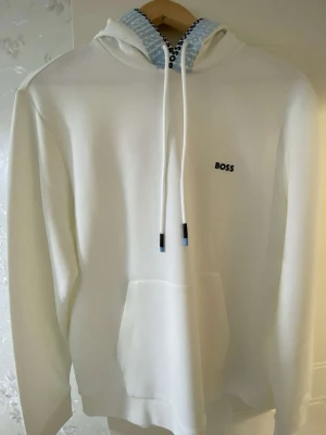 Vit hoodie från BOSS med logga - Snygg vit hoodie från BOSS med liten svart logga på bröstet och coolt mönster med BOSS-tryck på insidan av huvan. Klassisk känguruficka framtill och justerbara dragsnören. Perfekt för en clean och stilren look. Storlek L fast passar För M storlek är liten i passform.