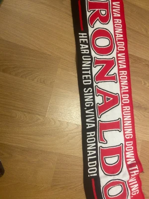 Röd och svart Ronaldo halsduk - Snygg halsduk med Ronaldo-tema i rött, svart och vitt. Stor text med 'Ronaldo' och detaljer som fransar i ändarna. Perfekt accessoar för fotbollsfans och United-supporters.