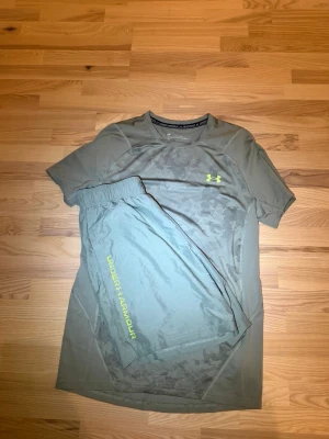 Under Armour träningsset grå/grön - Snyggt träningsset från Under Armour med t-shirt och shorts i grågrön färg. T-shirten har diskret mönster och logga i neongult på bröstet. Shortsen har elastisk midja och Under Armour-tryck längs benet. Perfekt för gym eller löpning.