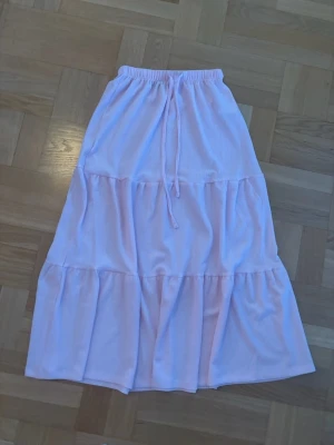 Ljusrosa volangkjol med knytband - Säljer en ljusrosa långkjol med volanger och elastisk midja med knytband. Kjolen har en luftig och flowy passform, perfekt för varma dagar. Materialet känns mjukt och bekvämt mot huden.