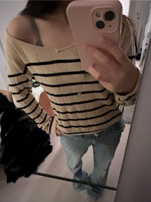 Beige randig tröja  - Trendig beige tröja med svarta horisontella ränder och kan klä som off shoulder-design. Långärmad och i mjukt, ribbat material som ger en avslappnad och snygg look. Perfekt att styla med jeans för en casual vibe. 