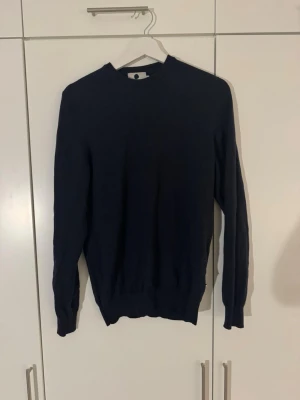 Nn07 stickad tröja - En enkel och stilren mörkblå stickad tröja med rund halsringning och långa ärmar. Tröjan har ribbade muddar vid ärmslut och nederkant, perfekt för lager på lager och en clean look.