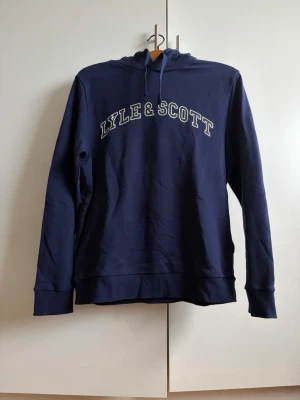 Marinblå hoodie från Lyle & Scott - Snygg marinblå hoodie från Lyle & Scott med stor logga i vitt på bröstet. Klassisk modell med huva och dragsnören, ribbade muddar vid ärmslut och nederkant. Tillverkad i mjukt bomullsmaterial som är skönt att ha på sig.
