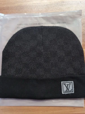 Svart mössa från Louis Vuitton - Snygg svart mössa från Louis Vuitton med diskret LV-logga framtill. Mössan har en uppvikt kant och är tillverkad i ett mjukt, stickat material som känns lyxigt och stilrent. Perfekt accessoar för dig som vill ha en exklusiv touch på din outfit.