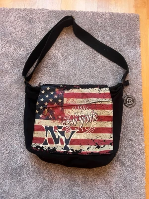 Axelväska Robin Ruth - Cool svart axelväska från Rubin Ruth med stort lock prytt av amerikanska flaggan och texten 'New York'. Väskan har justerbar axelrem och är tillverkad i tygmaterial. Perfekt för dig som gillar streetstyle och vill sticka ut med en unik design.