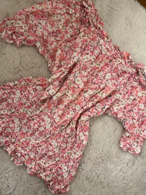 Blommig rosa blus - Supersöt blus med blommigt mönster i rosa, vitt och inslag av grönt. Klänningen har offshoulder-modell med volangdetaljer och smockad nederdel för en skön och trendig passform. Perfekt för sommarens alla tillfällen.