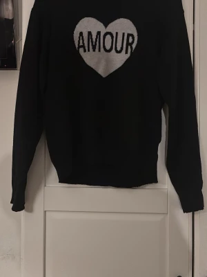 Svart stickad tröja med hjärta - Svart stickad tröja med ett stort grått hjärta och texten 'AMOUR' på bröstet. Tröjan har långa ärmar och ribbade muddar vid ärmslut och nederkant. Perfekt för dig som gillar en enkel men ändå statement look. En liten fläck på baksidan av ärmen.