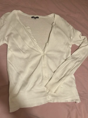 Vit basic kofta med knappar - En enkel och stilren vit kofta med långa ärmar och knäppning framtill. Koftan har en rundad halsringning och är tillverkad i ett mjukt, tunt material som passar perfekt till lager på lager. Snygg att matcha med jeans eller kjol. Passar även s