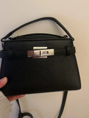 Svart handväska med silverdetaljer -  svart handväska i skinnimitation med struktur, topphandtag och justerbar axelrem. Väskan har en snygg silverfärgad låsdetalj framtill och minimalistisk design. Köparen står för frakten ☺️