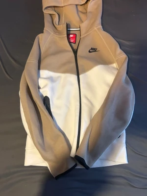Nike tech  beige och vit hoodie - Snygg hoodie från Nike i beige och vitt med dragkedja framtill och svart Nike-logga på bröstet. Hoodien har huva, långa ärmar och kontrasterande svarta detaljer vid dragkedjan och fickorna. Tillverkad i mjukt och bekvämt material, perfekt för en avslappnad stil.