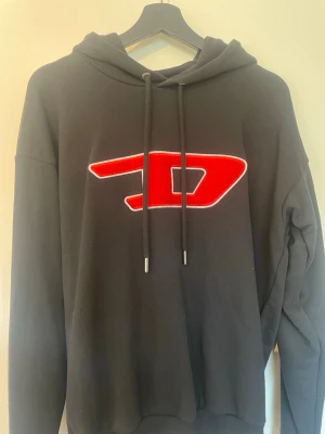 Svart hoodie från Diesel med röd logga - Svart hoodie från Diesel med stor röd och vit logga på bröstet. Klassisk huva med dragsko och långa ärmar. Tillverkad i mjukt bomullsmaterial som känns skönt mot huden. Perfekt för dig som vill ha en streetwear-look med attityd.