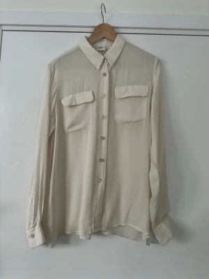 Vit skjortblus från exklusiva Caliban  - Elegant vit cremefärgad blus / skjorta från exklusiva Caliban. Storlek 46 om man vill ha tight fit, passar mig med storlek 42 om man vill ha lite luftigare fit. En ljus nyans med klassisk krage och knäppning framtill. Skjortan har långa ärmar med manschetter och två bröstfickor. Perfekt för både vardag och fest. Nypris 2200:-  Hämtas i Stockholm eller skickas mot porto. 