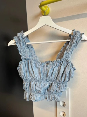 Blå linnetopp från Levi's med volang - Supersöt blå linnetopp från Levi's med breda axelband, volangdetaljer och knappar framtill. Toppen har smockad resår och broderade hål för en extra somrig vibe. Perfekt att styla med jeans eller kjol.