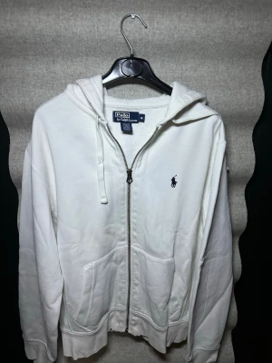 Vit hoodie från Polo Ralph Lauren - Klassisk vit hoodie från Polo Ralph Lauren med dragkedja framtill och broderad logga på bröstet. Tillverkad i mjuk bomull, har två fickor fram och justerbar huva med snören. Perfekt för en clean och stilren look.