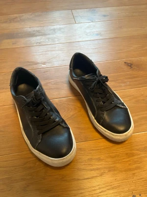 Svarta sneakers med vit sula - Stilrena svarta sneakers med vit platt sula och klassisk snörning. Skorna har en rund tå och är tillverkade i ett material som liknar skinn. Perfekta för en avslappnad och trendig look. Köpt på kings and queens i emporia för 900 kr