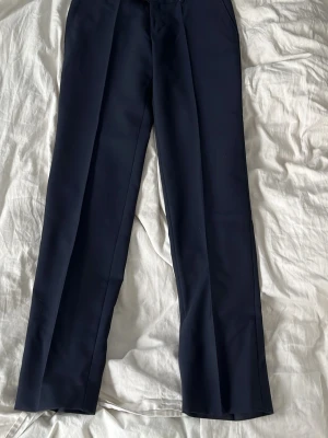 Mörkblå kostymbyxor med pressveck - Snygga mörkblå kostymbyxor med klassiskt rakt snitt och pressveck längs benen. Byxorna har en stilren design och är perfekta för en dressad look. Tillverkade i ett slätt material som ger en elegant känsla.