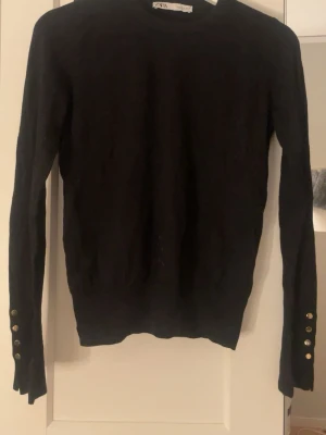 Svart stickad tröja från Zara - Svart stickad tröja från Zara med rund hals och långa ärmar. Snygga guldfärgade knappar vid ärmsluten ger en extra detalj. Perfekt att styla med jeans eller kjol för en trendig look.