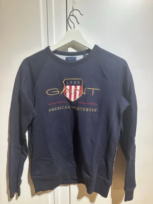 Mörkblå sweatshirt från Gant - Mörkblå sweatshirt från Gant med broderad logga och texten 'American Sportswear' framtill. Tröjan har rund halsringning, långa ärmar och ribbade muddar. Klassisk och sportig stil med rödvit sköld på bröstet. Tveka inte att skriva vid frågor eller funderingar 