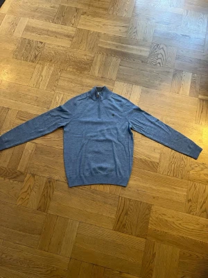 Blå halvzip tröja från Morris - En snygg och aldrig använd stickad tröja i blå/grå ton med half-zip och hög krage från Morris. Klassisk och tidlös design som funkar perfekt till både vardag och mer uppklädda outfits. Mjuk och bekväm – perfekt lager-på-lager under jacka eller kappa.