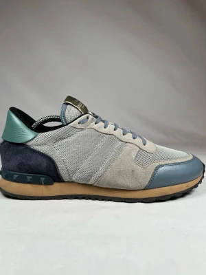 Valentino Rockrunner sneakers grå/blå - Snygga Valentino Rockrunner sneakers i grått och blått med detaljer i mocka, mesh och skinn. Skorna har klassisk snörning, rund tå och en beige sula med nitar på hälen. Perfekta för dig som vill ha en unik och trendig look.