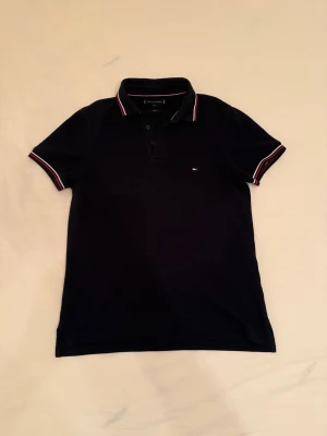 Tommy Hilfiger Piké - Klassisk marinblå pikétröja från Tommy Hilfiger med korta ärmar och krage. Små röda och vita detaljer på krage och ärmslut samt diskret logga på bröstet. Tillverkad i mjuk bomull för en bekväm känsla och normal passform.