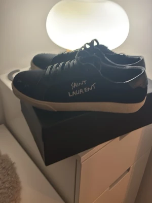 Svarta Saint Laurent sneakers - Svarta sneakers från Saint Laurent med vit sula och handskriven logga på sidan. Klassisk låg modell med snörning och rund tå. Tillverkade i skinn med stilren design som passar till många outfits.