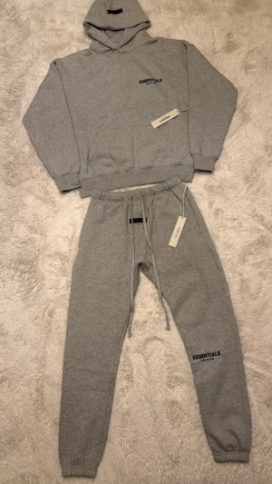 Grå Essentials Fear of God hoodie och mjukisbyxor - Snygg grå hoodie och mjukisbyxor från Essentials Fear of God med svart logga på bröstet och ryggen och under knät. Klassisk passform med huva och ribbade muddar. Perfekt för en avslappnad streetwear-look. Helt oanvänd och Storlek S.