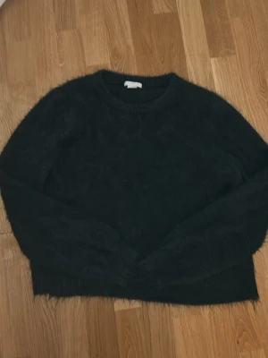 Mörkgrön fluffig stickad tröja H&M - Mysig mörkgrön stickad tröja från H&M med fluffig yta. Tröjan har rund halsringning och långa ärmar, perfekt för kyliga dagar. Enkel och stilren design som passar till många outfits.
