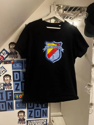 Blue saints - Svart t-shirt med ett stort färgglatt Blue saints emblem på bröstet. Skölden har blå, gul och röd färg och texten 'Blue Shirts' ovanför. Klassisk passform och rund halsringning. Perfekt för dig som gillar supporterstil.
