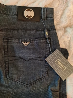 Svarta jeans från Giorgio Armani - Snygga svarta jeans från Giorgio Armani med klassisk femficksdesign och silverfärgad logga på bakfickan. Jeansen har raka ben och är tillverkade i ett mjukt bomullsmaterial med diskret struktur. Perfekta för dig som gillar stilrena och exklusiva plagg.