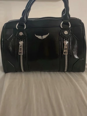 Svart handväska med vingar och dragkedjor - DEN ÄR INTE ÄKTA har få skador vid självaste märket och på sidan av men annars jätte fin Cool svart handväska i glansigt material med två silvriga dragkedjor framtill och ett emblem med glittrande vingar. Väskan har dubbla handtag och detaljer i silverfärg. Perfekt för dig som gillar edgy stil. 