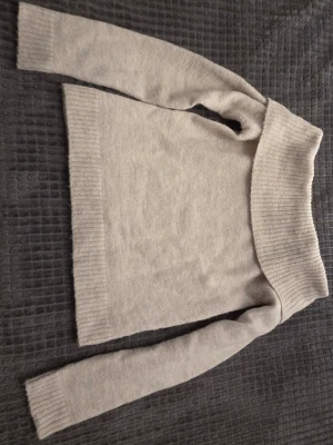 Beige off shoulder stickad tröja från Gina Tricot - Mysig beige stickad tröja med bred ribbad off shoulder-krage och långa ärmar. Tröjan har ribbade muddar vid ärmslut och nederkant, vilket ger en snygg och avslappnad look. Perfekt för dig som gillar en trendig och bekväm stil.