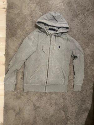 Grå zip hoodie från Polo Ralph Lauren - Snygg grå hoodie med dragkedja från Polo Ralph Lauren. Klassisk huva med vita snören och två fickor framtill. Ikoniska mörkblå loggan broderad på bröstet. Tillverkad i mjuk bomullsblandning som är skön att ha på sig. Perfekt till chill eller streetwear.
