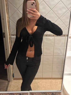 Svart långärmad knytblus - Trendig svart långärmad topp med djup v-ringning och knyt framtill. Snygg croppad modell som passar perfekt till höga jeans eller kjol. Tunt och stretchigt material som sitter skönt på kroppen.