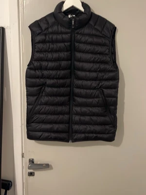 Svart quiltad väst med dragkedja - Stilren svart väst med quiltat mönster och hög krage. Västen har två snedställda fickor med dragkedja och stängs med en hel dragkedja framtill. Perfekt att slänga över en hoodie eller tröja för extra värme.