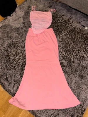 Ljusrosa långklänning med smala axelband - Snygg ljusrosa långklänning med smala axelband och figurnära passform upptill. Klänningen har en mjuk, utsvängd nederdel och är tillverkad i ett tunt, stretchigt material. Perfekt för dig som gillar en feminin och enkel stil.