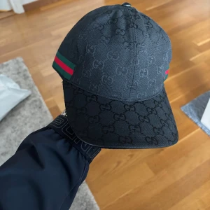 Svart keps från Gucci med GG-mönster - Snygg svart keps från Gucci med klassiskt GG-monogram över hela kepsen. Har gröna och röda ränder på sidorna och justerbar rem baktill. Tillverkad i bomullsmix och har en stilren, sportig look som sticker ut. Helt ny och oanvänd i perfekt skick