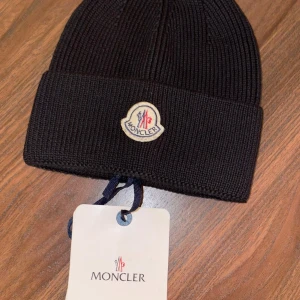 Svart ribbad mössa från Moncler - Snygg svart ribbstickad mössa från Moncler med klassisk uppvikt kant och broderad logotyp framtill. Perfekt accessoar för kyliga dagar och ger en stilren look. Tillverkad i mjukt stickat material för extra komfort.