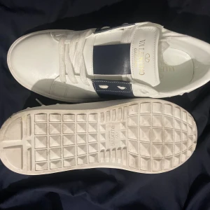 Valentino Garavani Open sneakers vit/blå - Säljer ett par snygga Valentino Garavani Open sneakers i vitt skinn med bred marinblå rem över mitten och coola nitar på sulkanten. Skorna har rund tå, platt sula och klassisk snörning. Perfekt för dig som gillar exklusiv streetstyle och italiensk design.