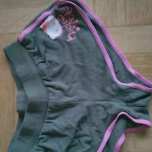 Hello Kitty shorts med rosa kant - Mörkgröna shorts med rosa kant och Hello Kitty-tryck framtill. Söt design med kontrasterande färger och en liten rosett på trycket. Perfekt för dig som gillar kawaii och vill ha något unikt i garderoben. Mjukt material som känns skönt mot huden.