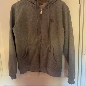Grå hoodie från Burberry med dragkedja - Snygg grå hoodie från Burberry med huva och snörning, hel dragkedja framtill och två stora fickor. Mjuk bomullskvalitet och diskret Burberry-logga på bröstet. Perfekt för chill dagar och enkel att matcha med jeans eller joggers.
