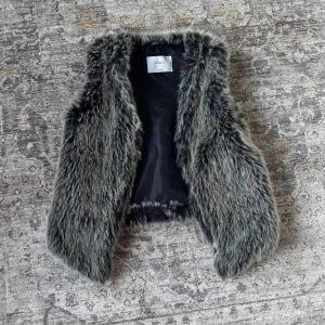 Grå pälsväst från ONLY - Snygg grå pälsväst från ONLY med fluffig och mjuk känsla. Västen har en mörkgrå ton med naturliga skiftningar och är fodrad med svart tyg på insidan. Perfekt att slänga över en hoodie eller långärmad tröja för extra edge.