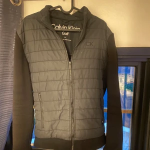 Cardigan jacka från Calvin Klein - Svart quiltad jacka från Calvin Klein Golf med dragkedja framtill och diskret CK-logga på bröstet. Jackan har vadderad framsida och axelparti, samt ärmar och rygg i mjuk fleece. Perfekt för dig som gillar sportig och stilren look. Nypris 1100kr!