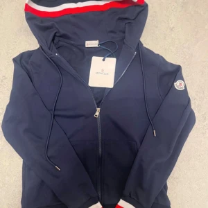 Mörkblå Moncler hoodie med huva - Snygg mörkblå hoodie från Moncler med dragkedja, huva och snörning. Huvan har röda och vita ränder och ärmarna har ribbade muddar med röd och vit detalj. Klassisk Moncler-logga på ärmen. Perfekt för en chill och sportig look.